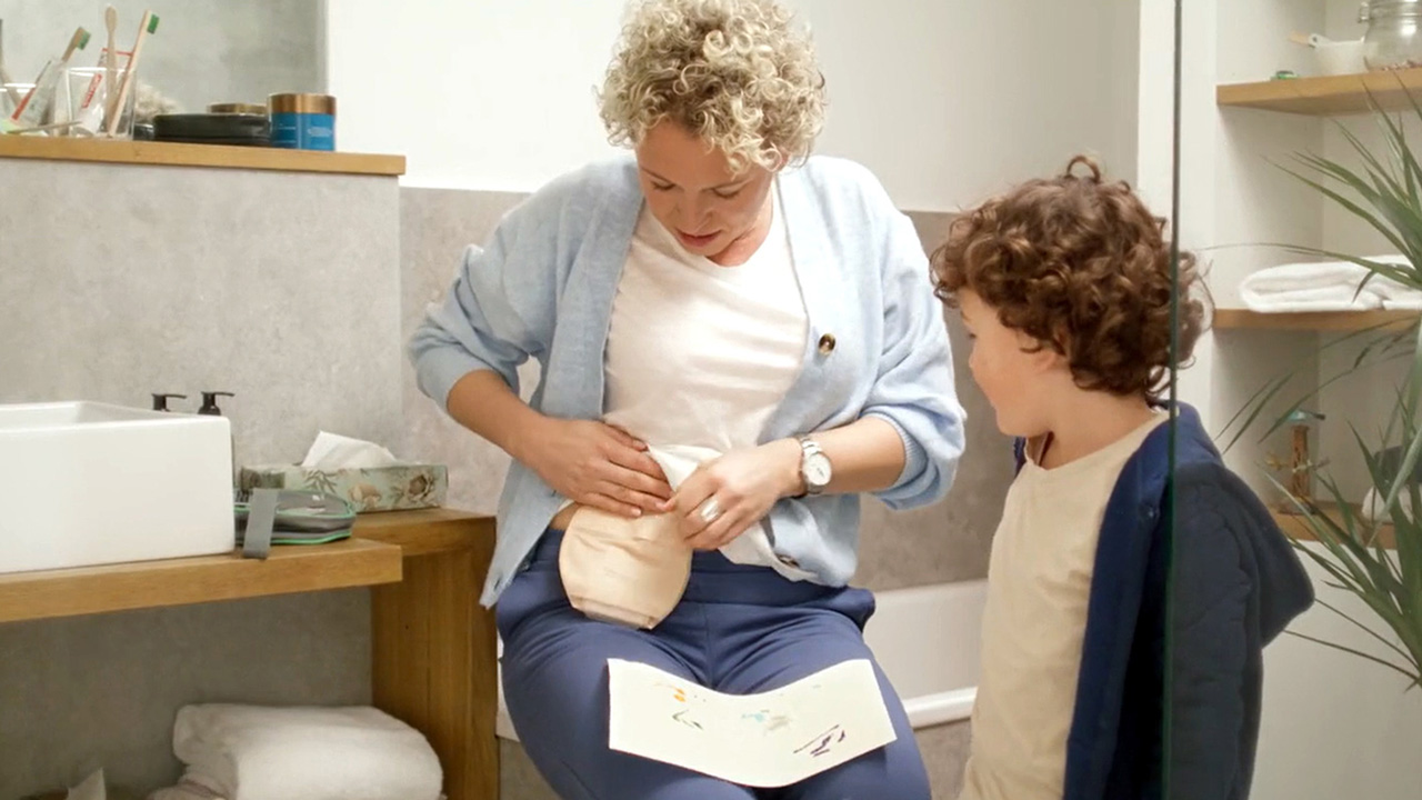 A woman in a white shirt sitting in the bathroom with a ostomy bag on her stomach and a manual on her knees. A child is standing next to her and looking on her ostomy bag. 