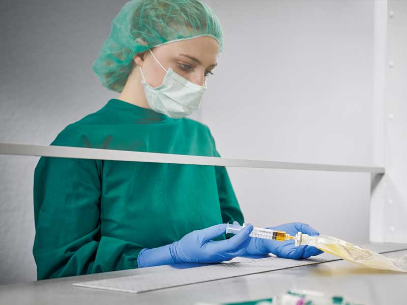 A female nurse dressed in sterile uniform with a face mask preparing parenteral nutrition in a sterile environment injecting a liquid into a nutrition bag. Various medical supplies are visible in the foreground.