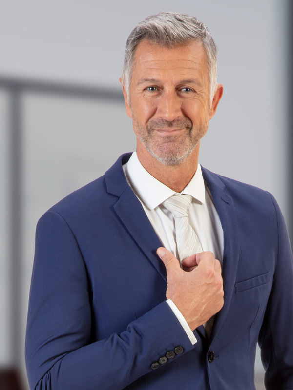 A middle-aged man dressed in a formal suit and tie, smiling warmly at the camera