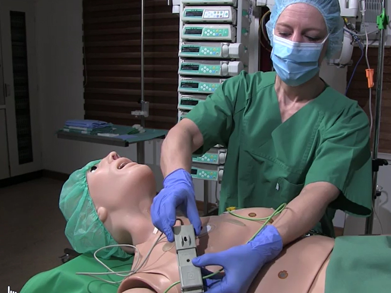 A healthcare specialist dressed in a green lab coat is testing a medical device on a mannequin patient lying on an examination table