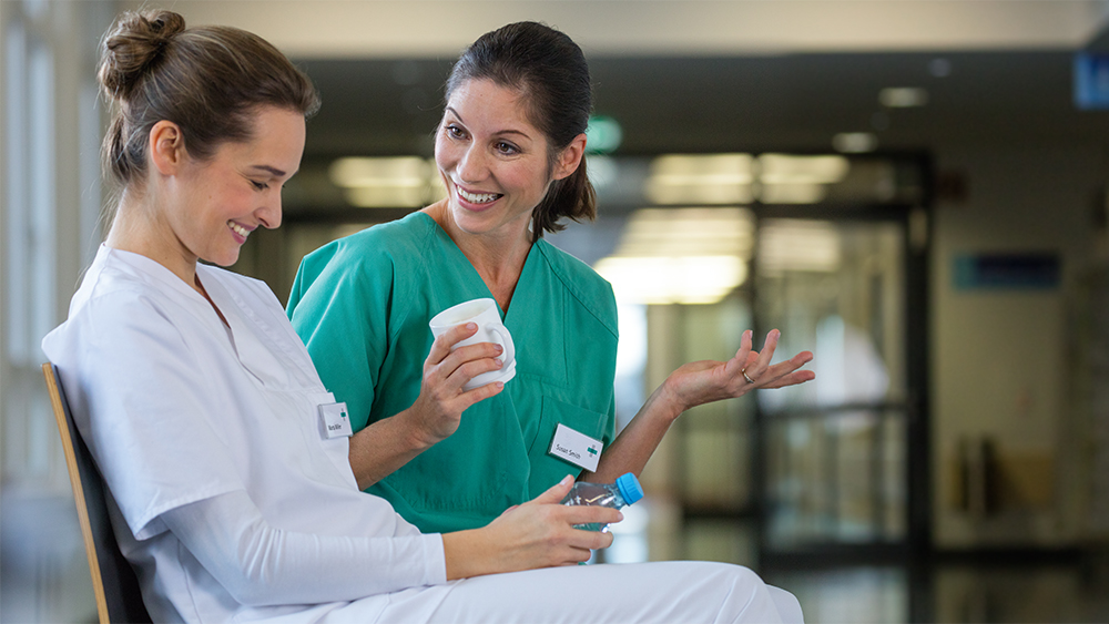 Two female nurses sitting in the hospital corridor taking their break drinking some coffee and water. Both are smiling while talking.  