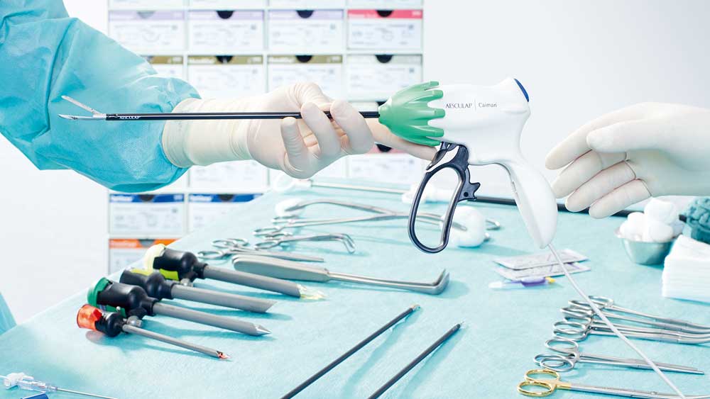 A detail of a minimally invasive surgical situation: one person in surgical clothes hands another a Caiman; in the background there is a table with surgical instruments neatly arranged for a surgery