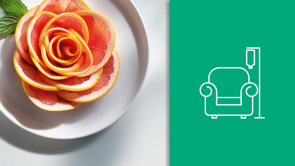 Grapefruit slices arranged in a flower shape on a plate. On a green background on the right, an icon of a chair with an infusion IV stand and hanging infusion bottle on the side representing home parenteral nutrition.
